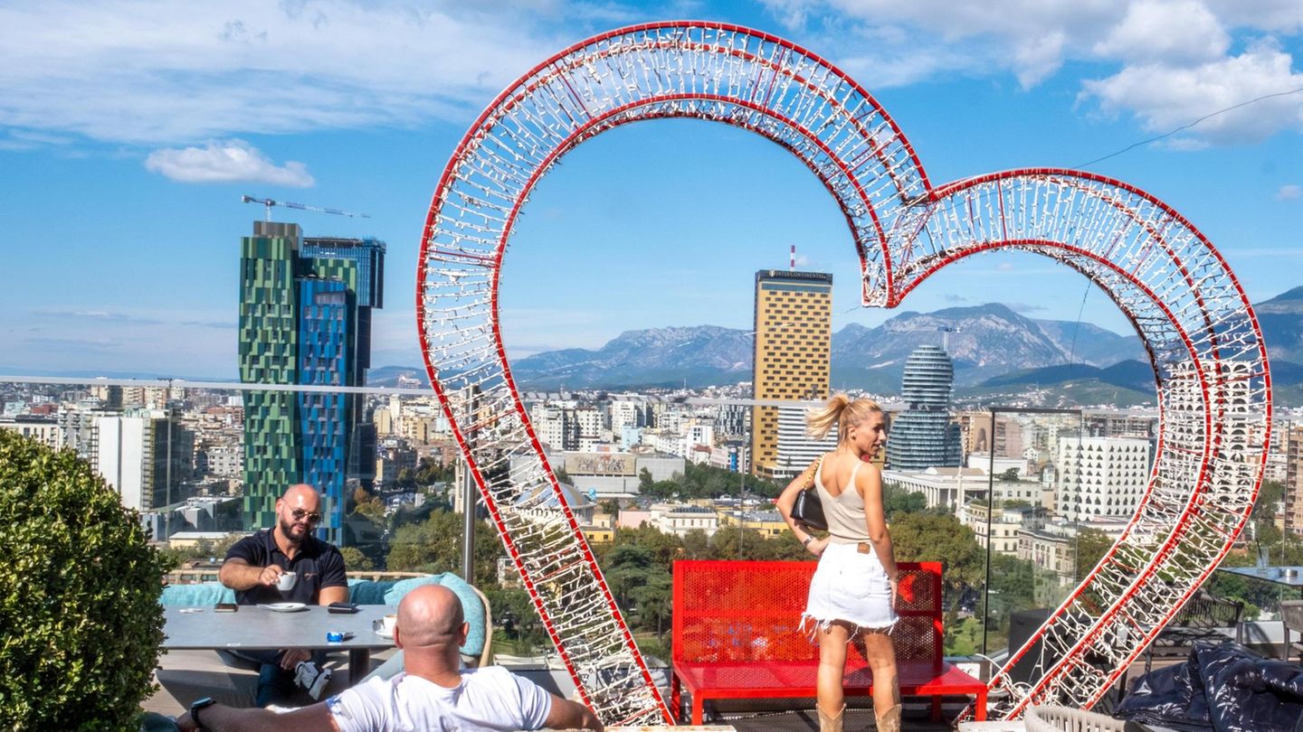 Eine Frau posiert vor einem Herzrahmen, im Hintergrund die Stadt Tirana