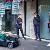 Ein Kleinkind sitzt in einem Spielzeugauto, drei junge Männer stehen dabei