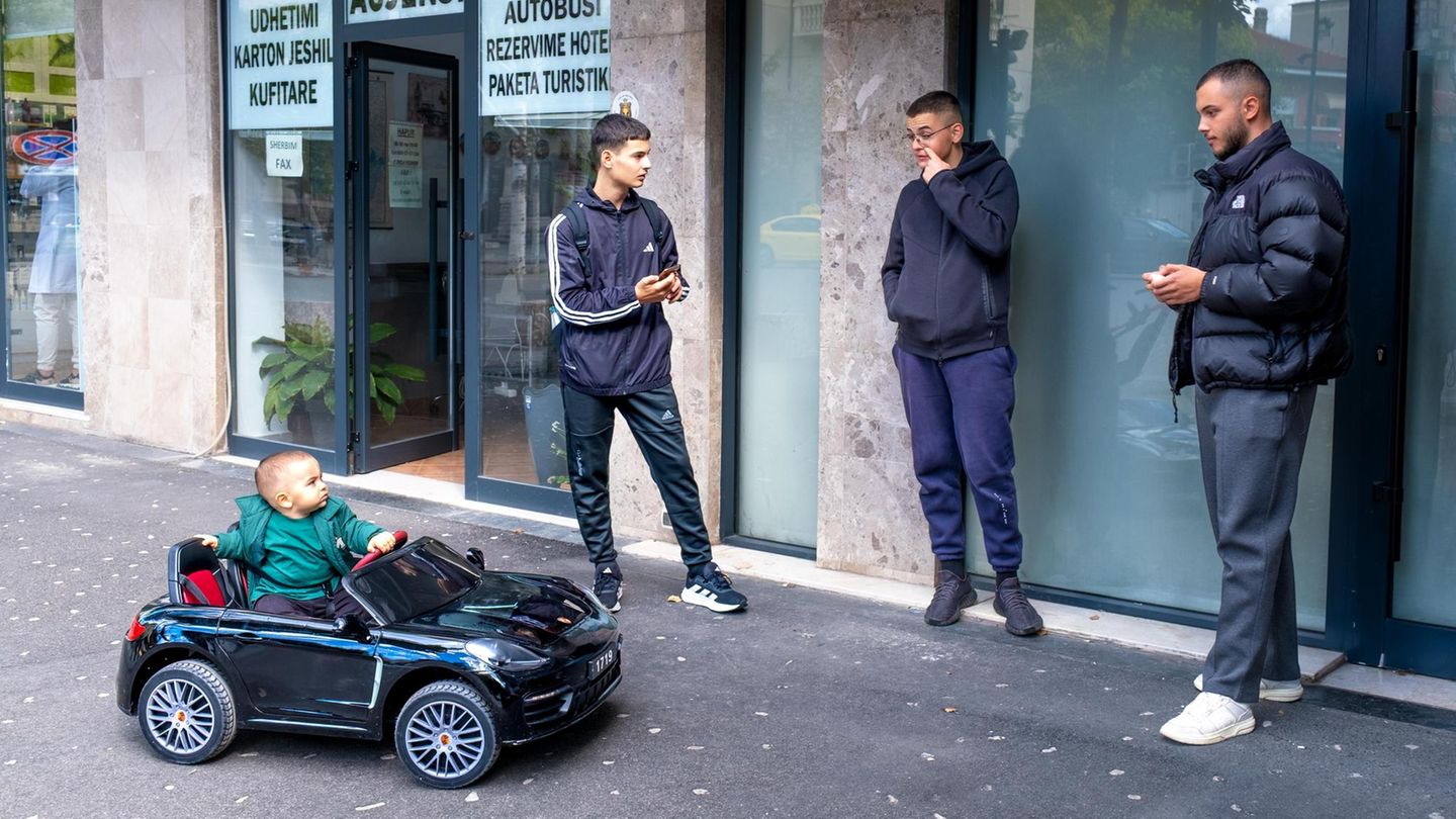 Ein Kleinkind sitzt in einem Spielzeugauto, drei junge Männer stehen dabei