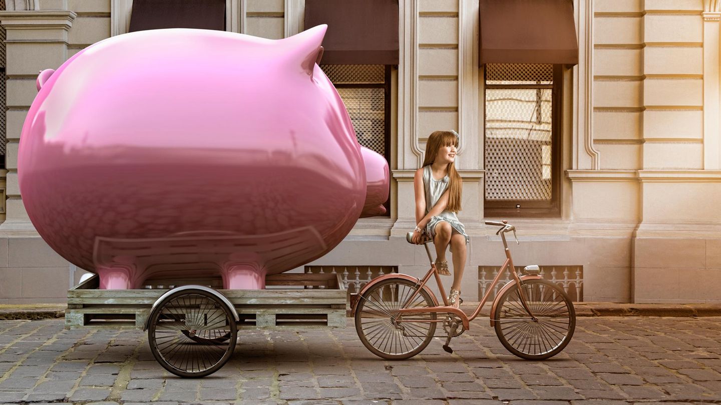 Ein Mädchen sitzt auf ihrem Fahrrad vor einem überdimensionalen Sparschwein, das auf einem Anhänger steht