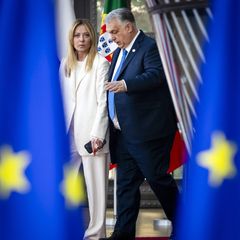 Meloni redet auf Orban ein, die Aufnahme vom EU-Gipfel ist durch zwei blaue EU-Fahnen hindurch fotografiert