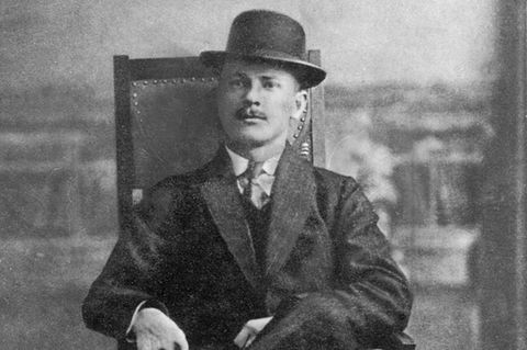 Petersen sitzt auf einem Holzstuhl, im Anzug und mit Melone