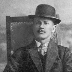 Petersen sitzt auf einem Holzstuhl, im Anzug und mit Melone