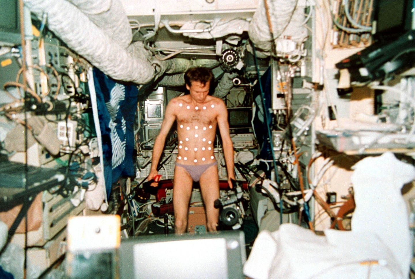 Ein Kosmonaut in Unterhose, den Oberkörper mit Punkten beklebt, führt ein Experiment durch.