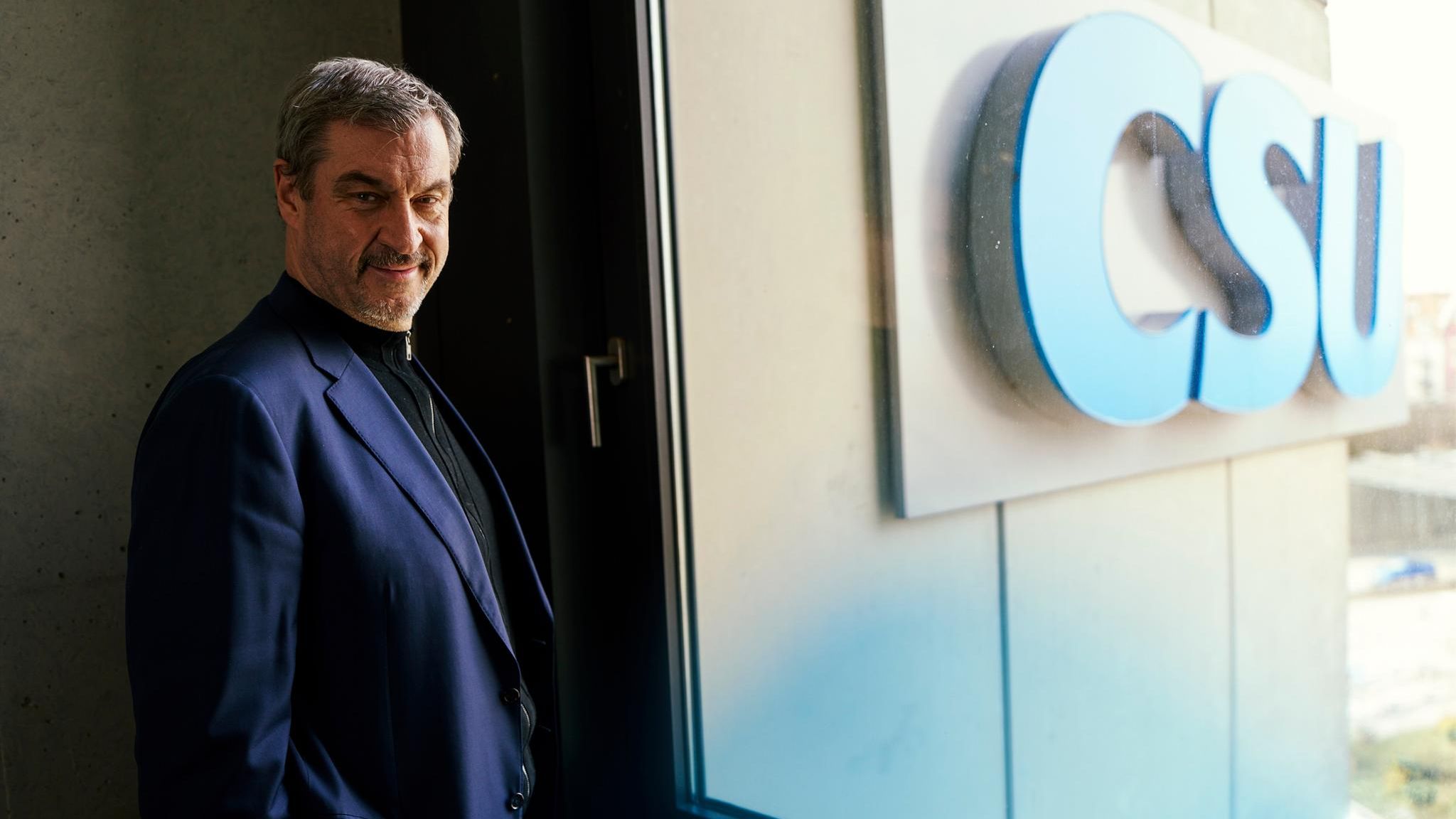 Markus Söder schaut in die Kamera, hinter einer Scheibe sieht man den CSU-Schriftzug