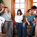 Fünf Menschen auf einem Sofa, die Paare links und rechts tuscheln, die Frau in der Mitte schaut genervt