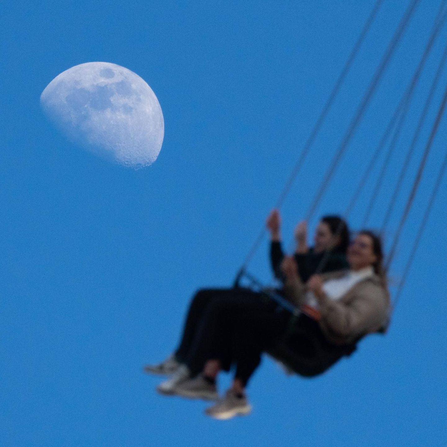 Zwei Frauen sitzen in einem Kettenkarussel, im Hintergrund ist gut der Mond zu sehen