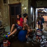 Kinder sitzen auf Regentonnen, Frauen stehen im Wasser
