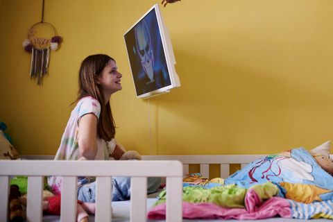 Das Mädchen Emmi sitzt auf eine Bett, nah vor einem Bildschirm. Sie leidet unter Kinderdemenz