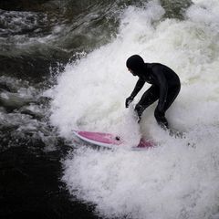 Ein Surfer in schwarzem Neopren surft auf pink-weißem Board die Eisbachwelle in München