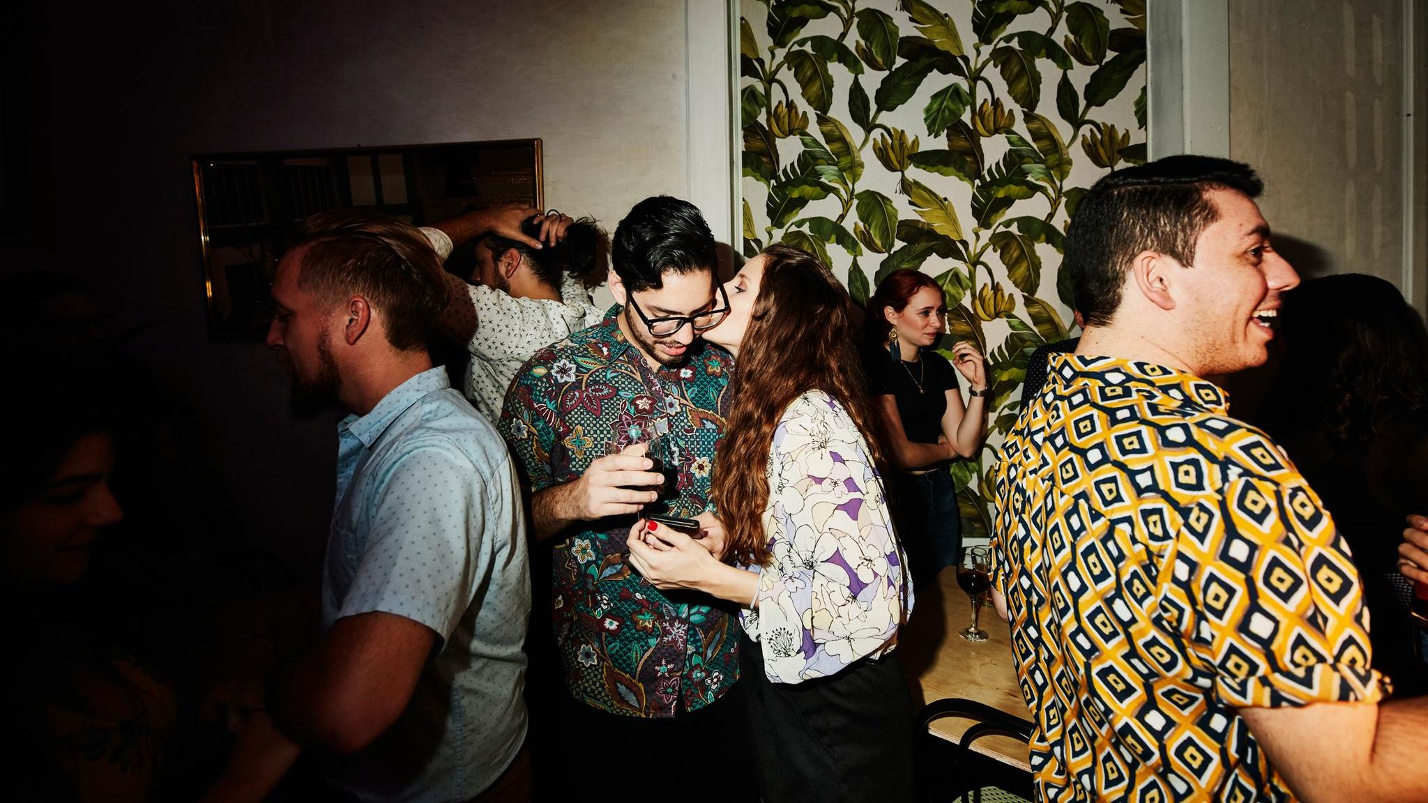 Partyszene: Eine Frau küsst einen Mann, andere Gäste stehen im Hintergrund. Ist es Liebe?