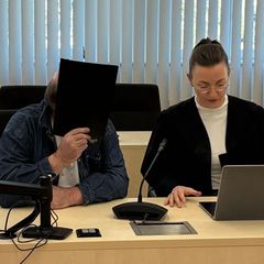 Der Angeklagte sitzt neben seiner Verteidigerin und hält sich eine Mappe vor das Gesicht