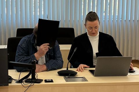 Der Angeklagte sitzt neben seiner Verteidigerin und hält sich eine Mappe vor das Gesicht