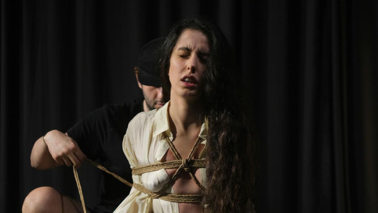 Seile schlingen sich um den Oberkörper einer schwarzhaarigen Frau. Hinter ihr kniet ein Mann. Sie praktizieren Shibari.