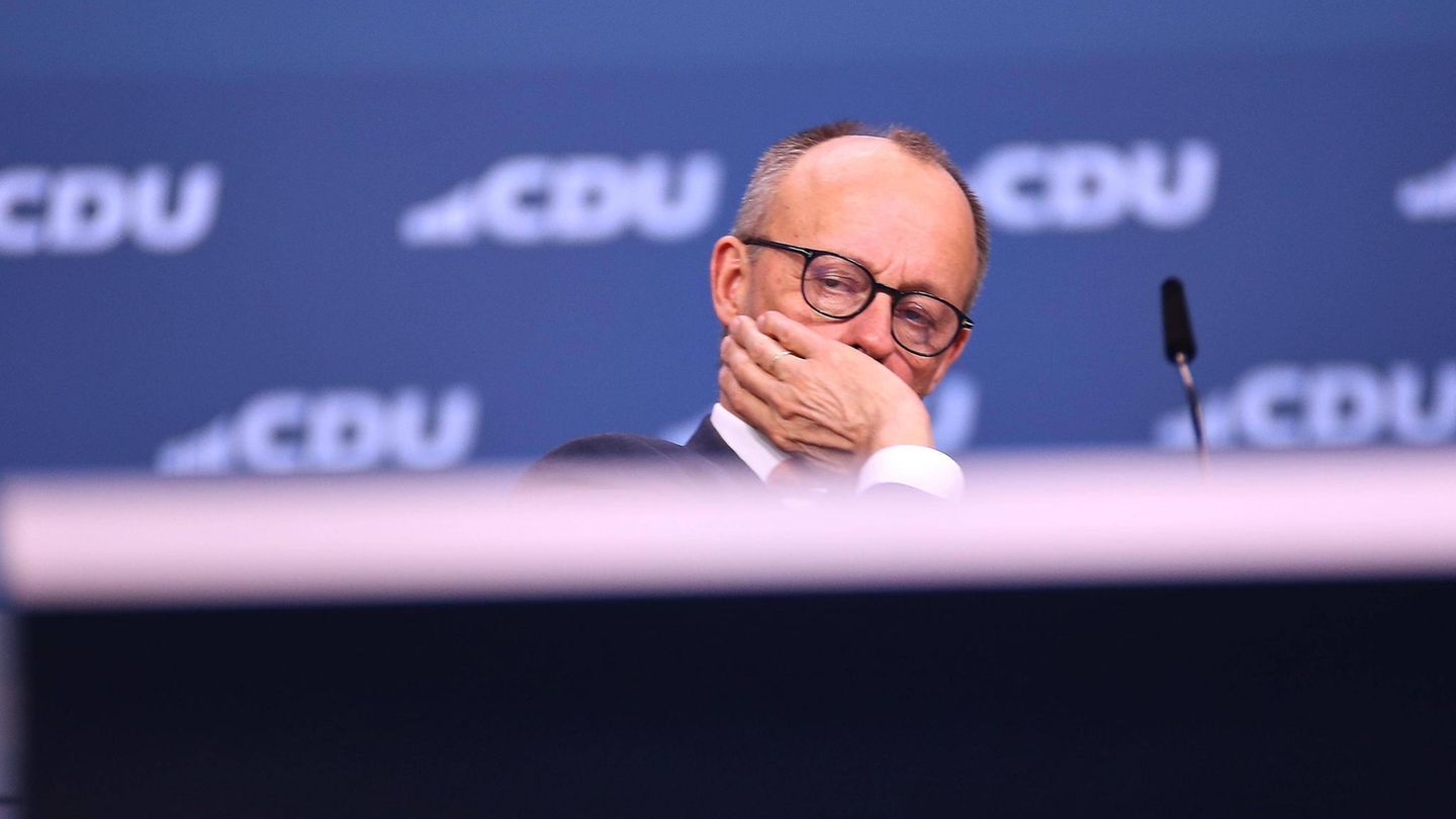 Friedrich Merz sitzt auf einer Bühne, die Hand vor dem Gesicht