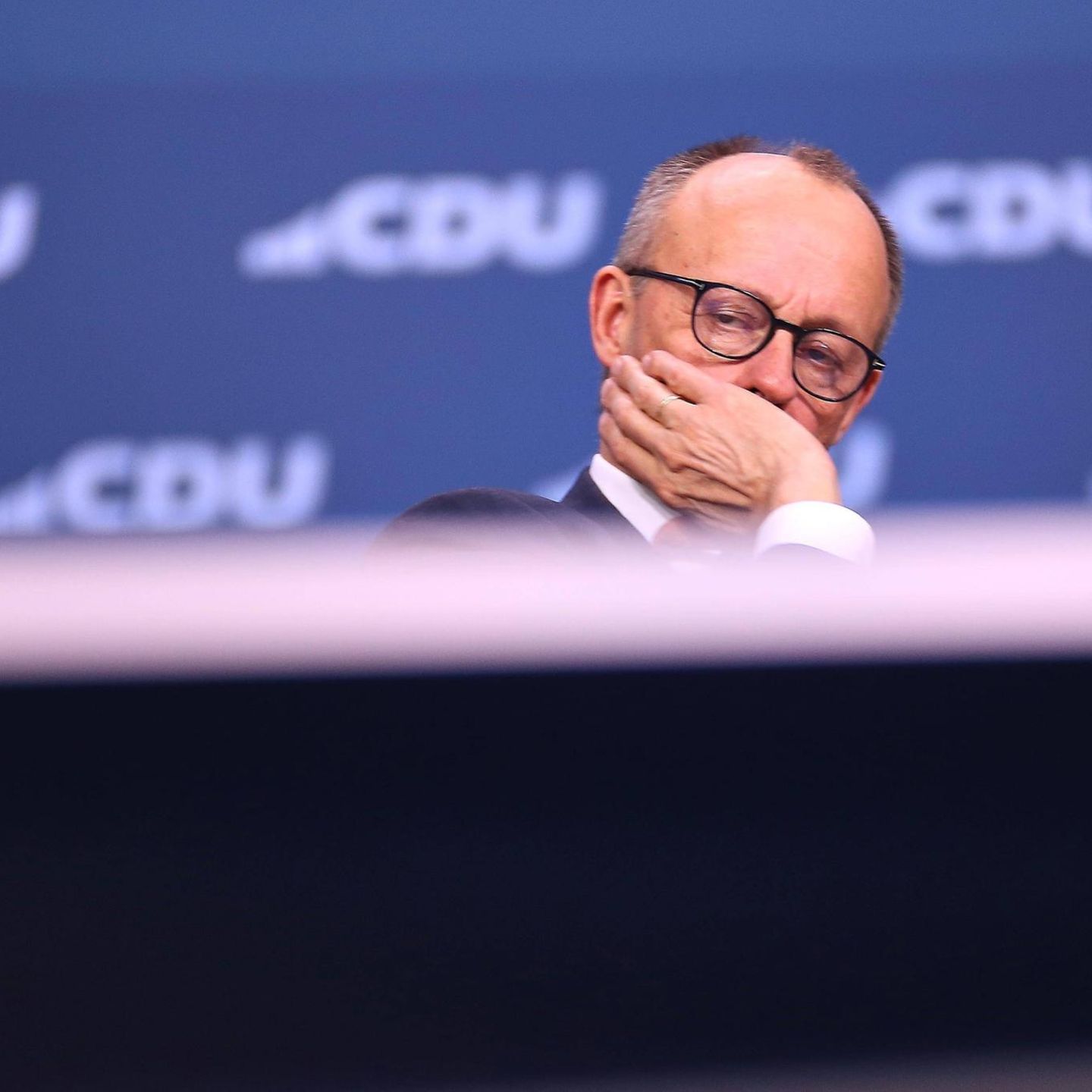 Friedrich Merz sitzt auf einer Bühne, die Hand vor dem Gesicht