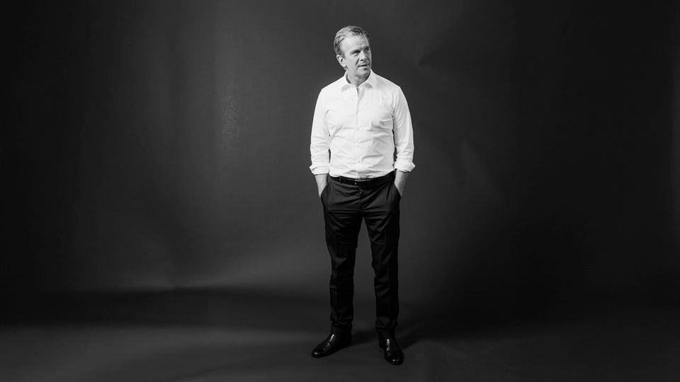 Markus Lanz steht vor einem  Fotohintergrund und schaut zur Seite