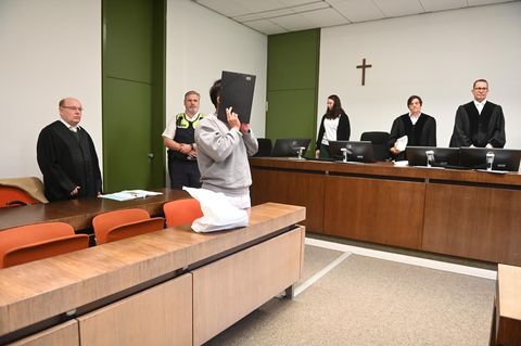 Der Angeklagte steht vor der Anklagebank und hält sich einen Aktenordner vors Gesicht