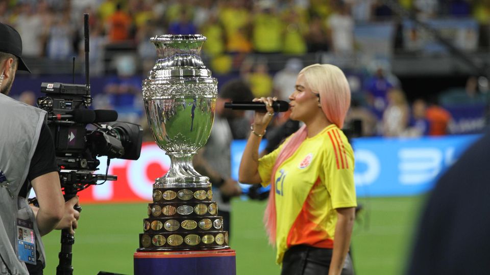 Karol G trägt ein kolumbianisches Fußball-Trikot, steht neben einem Pokal und singt in ein Mikro