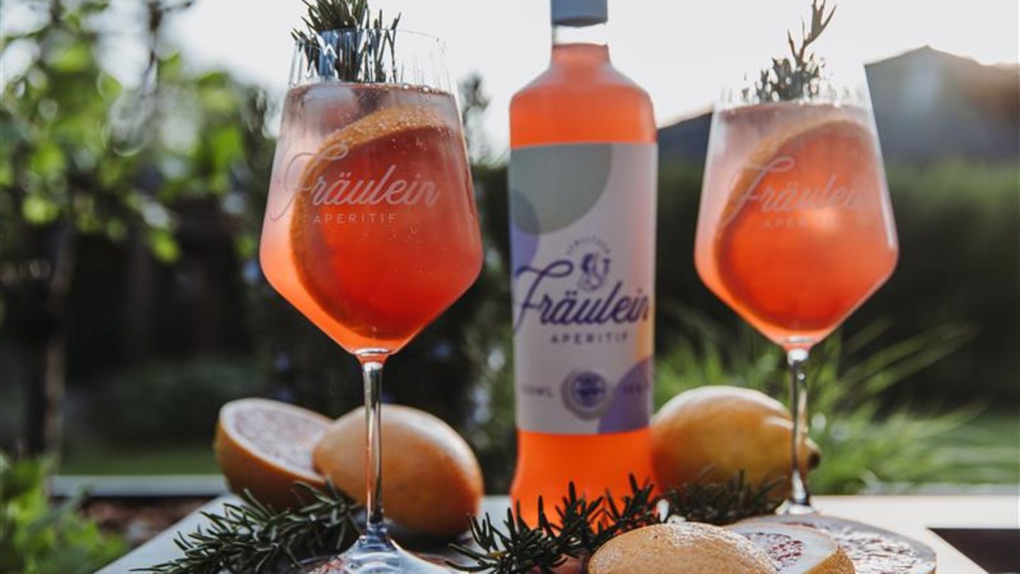 Eine Flasche mit orangener Flüssigkeit sowie zwei großbauchige Gläser mit gleichem Inhalt vor einer Berg-Kulisse im Hintergrund