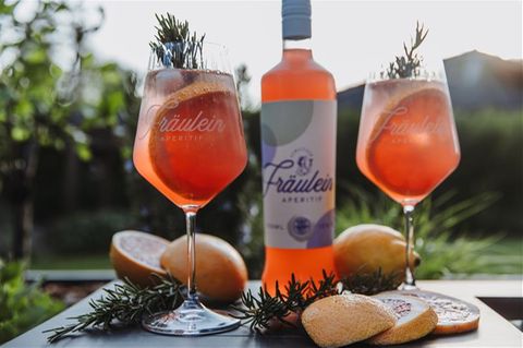 Eine Flasche mit orangener Flüssigkeit sowie zwei großbauchige Gläser mit gleichem Inhalt vor einer Berg-Kulisse im Hintergrund