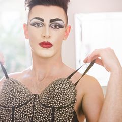 Fetisch Crossdressing: Ein geschminkter Mann hält eine Corsage vor seinen nackten Oberkörper