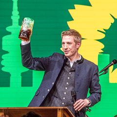Dominik Krause hebt am Rednerpult eine Maß Bier.