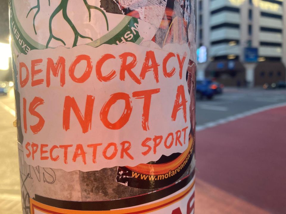 Aufkleber an einem Ampelmast mit dem Satz "Democracy is not a spectator sport"