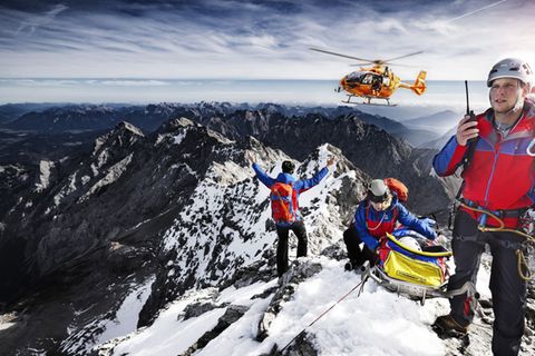 Auch die zweite Staffel der "Bergretter im Einsatz" zeigt Bergwachteinsätze in den bayerischen Alpen – hautnah und in beeindruckenden Bildern. Drei Angehörige der Bergwacht, die sich mit einer geretteten Person auf einem Berggipfel befinden, weisen einen anfliegenden Hubschrauber ein.