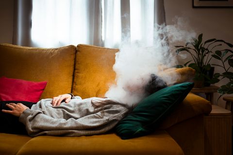 Nebel im Gehirn – Brainfog Symbolbild: Eine Person liegt auf einer gelben Couch, ihr Kopf ist von Nebel umhüllt