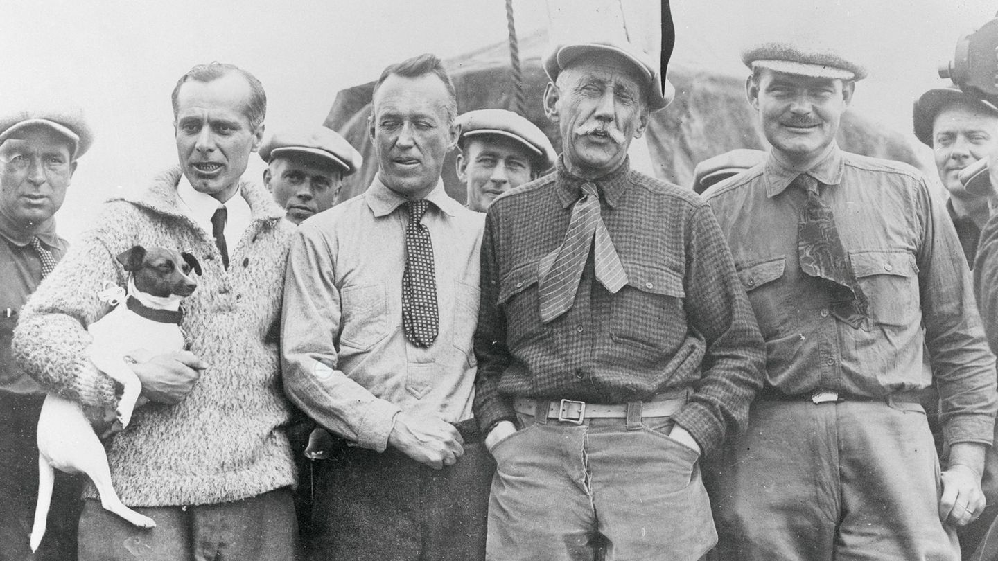Nobile, der seinen Hund auf dem Arm hält, steht neben Ellsworth und Amundsen sowie weiteren Männern der Crew.