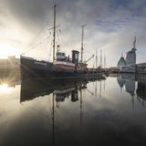 Ein Schiff im Hafen mit beeindruckender Spiegelung im Wasser bei Sonnenuntergang vor einer modernen städtischen Kulisse