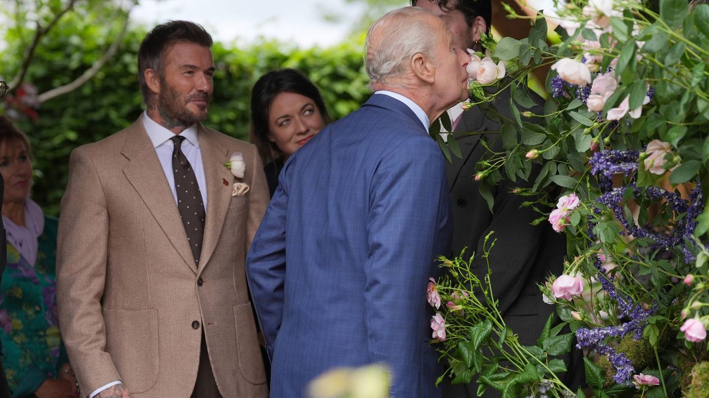 König Charles riecht an einer Blume, im Hintergrund steht David Beckham