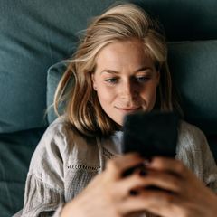 Eine Frau liegt mit dem SMartrphone auf dem Bett. Die Geburtenrate leidet