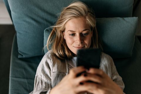 Eine Frau liegt mit dem SMartrphone auf dem Bett. Die Geburtenrate leidet