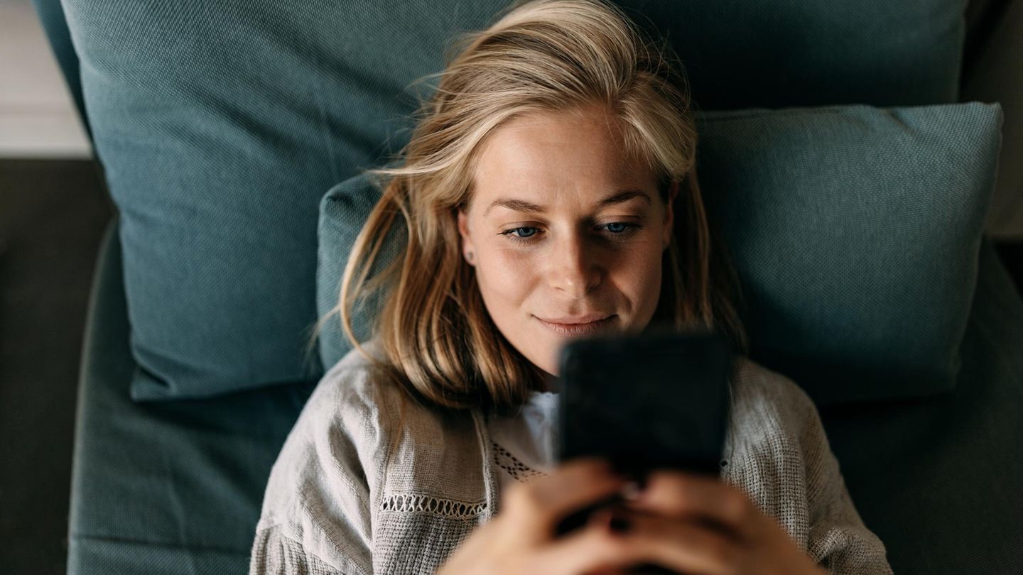 Eine Frau liegt mit dem SMartrphone auf dem Bett. Die Geburtenrate leidet