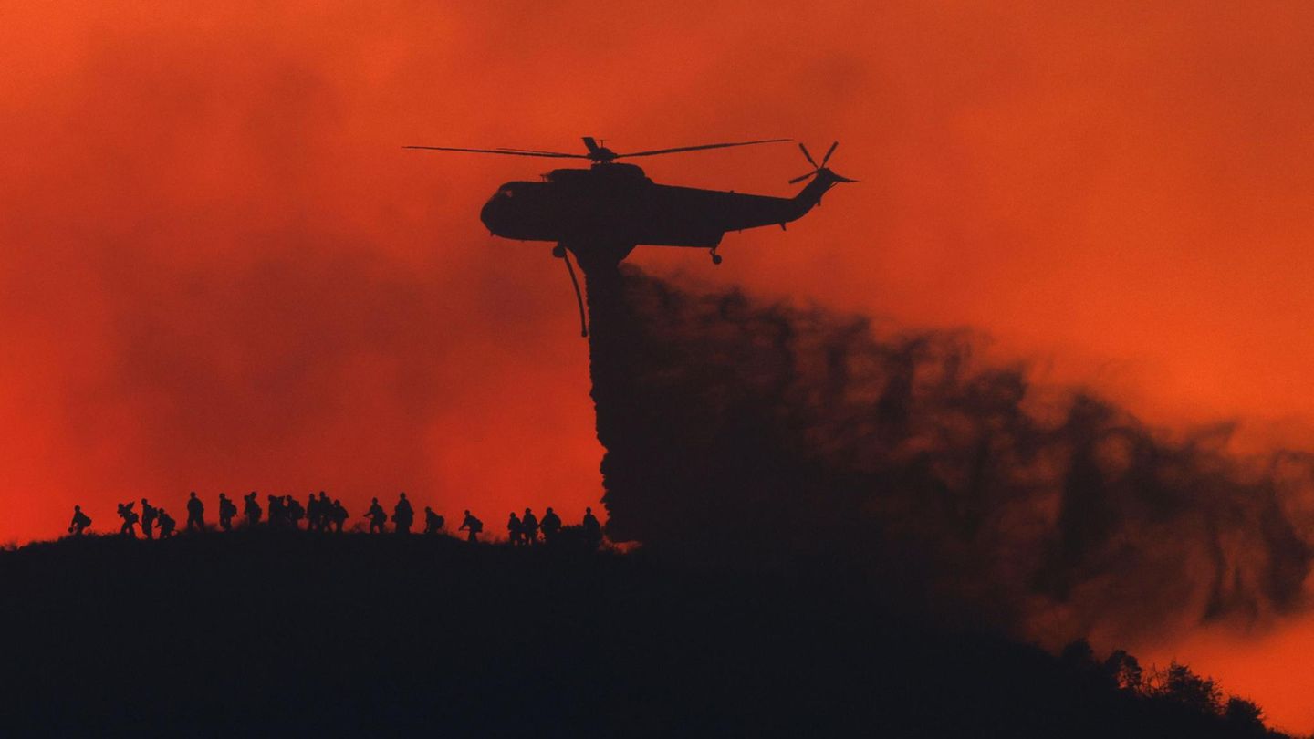 Ein Löschhelikopter wirft Wasser ab, während Feuerwehrleute unter ihm laufen