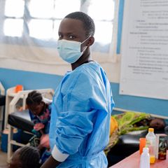 Ebola Virus: Ein Pfleger mit Maske blickt in die Kamera, im Hintergrund sind Menschen auf einem Krankenbett