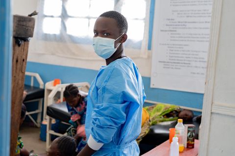 Ebola Virus: Ein Pfleger mit Maske blickt in die Kamera, im Hintergrund sind Menschen auf einem Krankenbett