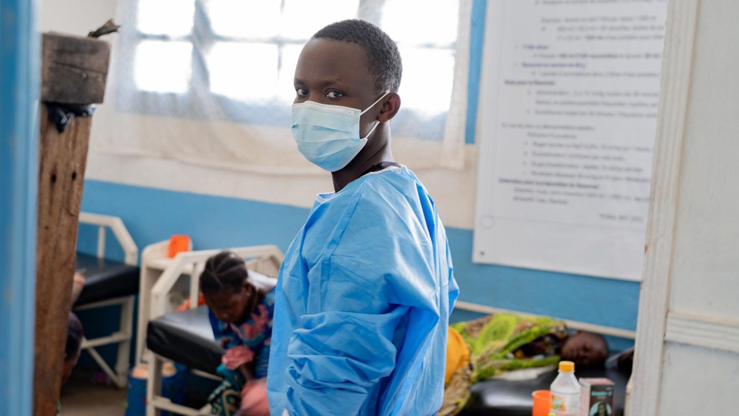 Ebola Virus: Ein Pfleger mit Maske blickt in die Kamera, im Hintergrund sind Menschen auf einem Krankenbett