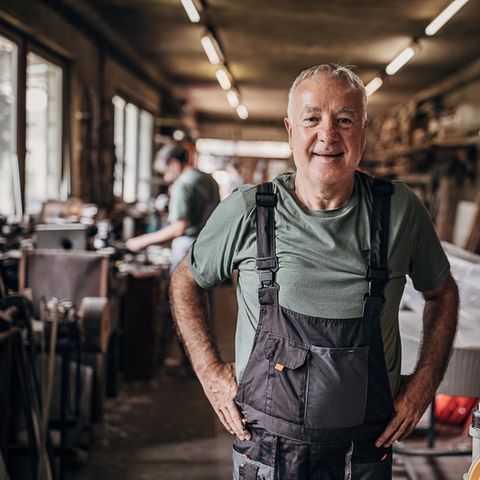 Älterer Mann in blauer Latzhose steht in der Werkstatt, ob er bis zur Rente mit 70 arbeitet?