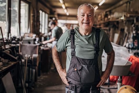 Älterer Mann in blauer Latzhose steht in der Werkstatt, ob er bis zur Rente mit 70 arbeitet?