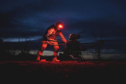 Ein Soldat bei Nacht, eine rote Lampe auf seinem Helm erleuchtet die Drohne, über die er sich beugt.