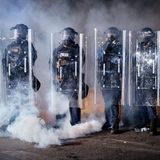 Sechs Polizisten stehen vermummt in der Reihe und haben Schutzschilder vor sich aufgebaut