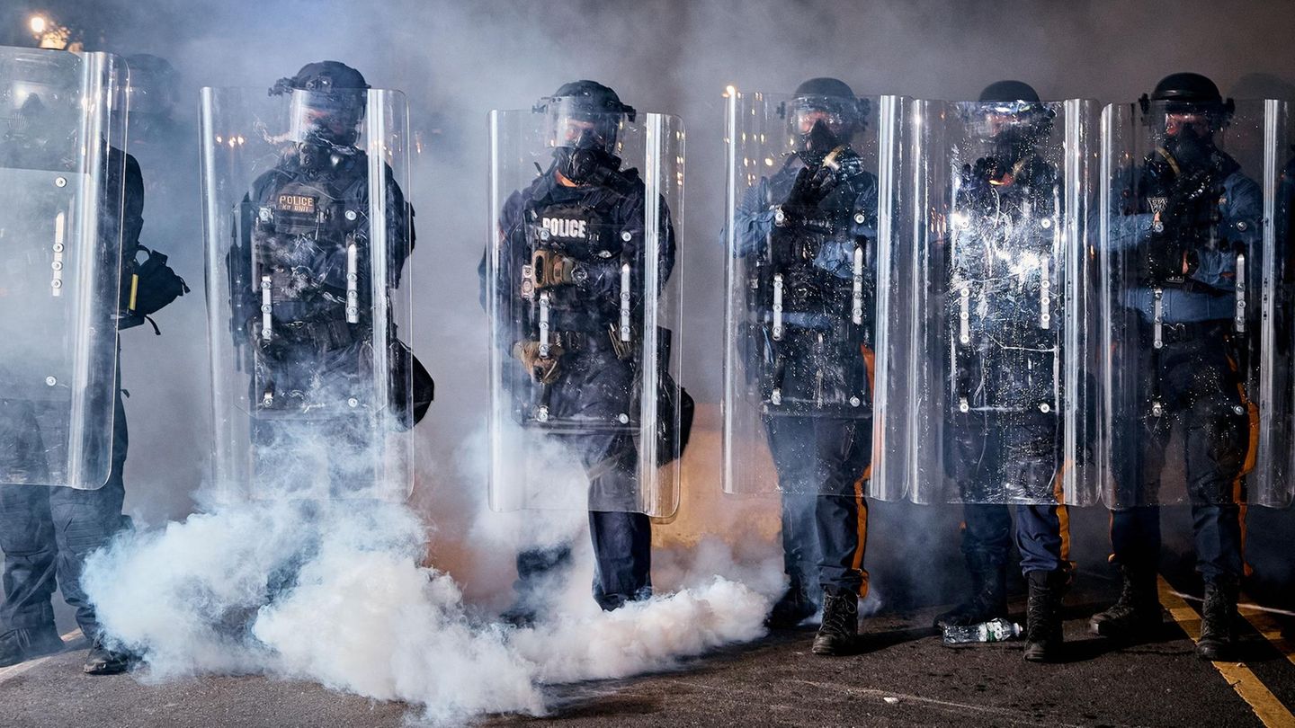 Sechs Polizisten stehen vermummt in der Reihe und haben Schutzschilder vor sich aufgebaut