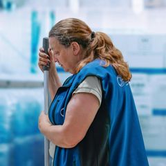 Eine Putzfrau in blauer Uniform hält die Stange eines Wischmopps fest – Szene des Films „Ich verstehe Ihren Unmut“