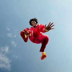 Eine Frau schwebt über der Kamera, sie lächelt. Im Hintergrund ist der Himmel zu sehen.