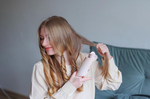 Lächelnde junge Frau, die zu Hause ihre Haare mit einem modernen Lockenwerkzeug