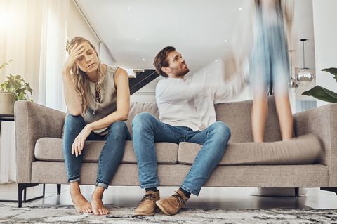 Zwei Eltern sitzen auf dem Sofa. Mutter fasst sich im Stress an den Kopf. Vater greift nach dem springendem Kind.