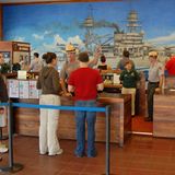 Pearl Harbor  Sie warten auf Einlass zum Freilichtmuseum, wo einst für die USA der Zweite Weltkrieg begann: Hawaiis Hauptattraktion ist die Gedenkstätte für die Opfer des Überraschungsangriffs. "This is not a drill - dieses ist keine Übung", lautete ein verzweifelter Funkspruch am Morgen des 7. Dezember 1941. Die militärische Niederlage bleibt beim Rundgang durch den öffentlichen Teil der Marinebasis stets gegenwärtig. Das aus Spenden finanzierte Visitor Center beherbergt einen Kinosaal und einen Souvenir Shop mit Dutzenden von DVD-Dokumentationenen und dem Reprint einer Tageszeitung, deren Zeile lautet: "War declared. 3000 Casualties in Jap Attack on Hawaii".